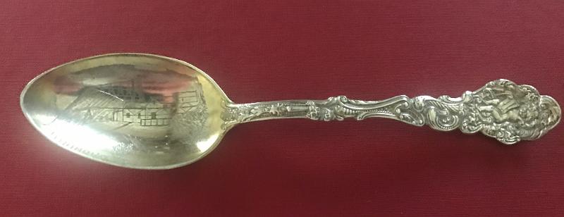 Souvenir Mining Spoon Oronogo No. 1 Mine, Oronogo,  MO.JPG - SOUVENIR MINING SPOON ORONOGO MINE NO 1 ORONOGO MO [Sterling silver spoon, 5 7/8 in. long, engraved mining scene in bowl, bowl marked OROGONO NO. 1, handle front in ornate Versailles pattern introduced in 1888 by Gorham;  It shows an angel holding a musical instrument, maybe pipes or a lyre; reverse is marked with the old lion-anchor-G  hallmark for Gorham, K in a square shape and Sterling; weight 30.9 gms. [Eight miles north of Joplin, Missouri stands the village of Oronogo, originally named Minersville when it sprang up in 1850. According to legend, a miner dug there with his pick and shovel and said, "It's ore here or no go," and so the name Oronogo stuck. Nearby is the site of the richest lead and zinc strike ever made in southwest Missouri. Tom Livingston first discovered the mine before the Civil War, but renegades stole his land while he was away fighting. After the war, they sold it to Granby Mining Company for $50. After almost 80 years of continuous operation, the mine produced $30 million worth of lead and zinc. The Oronogo Circle Mine No. 1, one of the few open-pit mines in the district, stretched 300 feet deep and 600 feet across. The 12-acre unroofed cavern was strip mined at three levels-150 feet, 240 feet, and 360 feet--a flurry of activity with men, trucks, and machinery going up and down the steep inclines. Numerous mills serviced the operation. The largest chunk of pure lead ever found in the district came from the Oronogo Circle. Two flat cars carried it to the World's Fair in Chicago in 1893. After the exhibit ended, the mammoth rock sold for $6,000. A deadly accident occurred at the mine in 1901 when a blast of dynamite detonated prematurely, killing 12 men and wrecking a $50,000 mill. Operations shut down for several months because miners refused to go back to work. Chicago capitalists ran the mine from 1906 to 1914, raking in profits of $3 million. They sold it to the Connecticut Mining Company for $500,000. The ground was so honeycombed with shafts and drifts at four levels that it was no longer safe to work underground. In an ill-fated endeavor to make mining the Circle safer, the new owners closed the old shafts and dismantled 20 small mills. They removed underground support pillars and stripped the entire tract down to 300 feet, using the largest steam shovels available. Then they buried dynamite at the former mill sites and ignited them all at once, causing a blast that almost blew Oronogo off the map. The ground shook so violently that a new mill, built on solid ground, fell into the pit and killed six men. The catastrophe cost the company more than $1 million. Connecticut Mining went out of business, and the mine reverted to its original owner, the Granby Mining Company. For many years, the tract lay idle, although investors made half-hearted attempts to resume strip mining. By the 1970s, the huge water-filled pit had become popular with scuba divers, who liked to explore its depths, yet even that turned out tragically, with the deaths of some of the divers.]